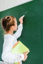 cute schoolkid holding chalk and books near green chalkboard .