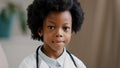 Cute serious african american kid girl in medical clothes dressed in white coat standing indoors posing looking at Royalty Free Stock Photo