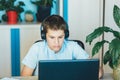 Cute serious boy in blue shirt sitting behind desk in his room next to laptop and study. Teenager in earphones makes homework,