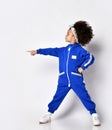 Cute serious dark-skinned mulatto kid girl in warm blue trendy sport jumpsuit and white sneakers stands pointing aside