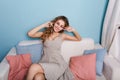 Cute girl with blond curly hair sits on sofa in studio with blue walls and colorful pillows. She wears light dress Royalty Free Stock Photo