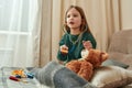 A cute small girl playing doctor using a toy stethoscope on her fluffy patient sitting on a sofa in a big well-lighted