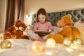 A cute small girl wearing pyjamas sitting alone cross-legged on a huge bed barefoot watching cartoons on a tablet and Royalty Free Stock Photo