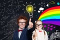 Cute smart kids showing thumb up against chalkboard background with light bulb, science formulas and arts pattern.