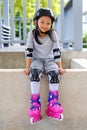 Cute smiling Asian kid girl in roller skates and protective gear sitting in the park