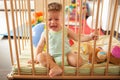 Sad crying cute baby looking through the wooden bars of his crib or playpen indoors in the nursery