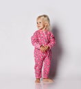 Smiling barefooted blonde baby kid girl in pink warm comfortable jumpsuit stands over grey wall background and looks at side