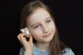 Cute smiling blonde girl holding seashell next to her ear and listening to the ocean on black background in studio Royalty Free Stock Photo