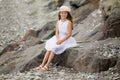 A cute smiling blonde girl 9 years old in a hat and white dress sits on the seashore Royalty Free Stock Photo