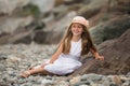 A cute smiling blonde girl 9 years old in a hat and white dress sits on the seashore Royalty Free Stock Photo