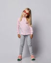 Cute blonde kid girl in stylish smoked heart-shaped sunglasses, turtleneck shirt sweater and pants is posing over gray Royalty Free Stock Photo
