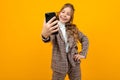 Cute smiling blondy girl in a classic jacket makes selfie on the phone in the studio with a yellow background