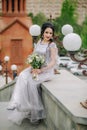 Cute smiling bridesmaid sitting on stone in front of the church, holding bridal bouquet and looking at camera Royalty Free Stock Photo