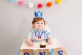 Cute smiling Caucasian baby boy in blue crown celebrating his first birthday at home. Child kid toddler sitting in a high chair Royalty Free Stock Photo