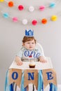 Cute smiling Caucasian baby boy in blue crown celebrating his first birthday at home. Child kid toddler sitting in high chair Royalty Free Stock Photo