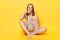 Cute smiling delighted young woman wearing striped swimming suit isolated on yellow background sitting holding her hat looking at Royalty Free Stock Photo