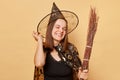 Cute smiling happy young woman wizard wearing witch costume holding in hand broom  over beige background looking at camera Royalty Free Stock Photo