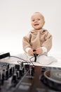 A cute smiling kid in a beige hoodie sits on the floor and plays with dj headphones and a dj mixing console. Music and