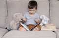 Cute smiling little baby boy reading book sitting on sofa with teddy bear toy and bunny rabbit at home. Royalty Free Stock Photo