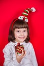 Cute smiling little girl in elf hat holding festive gift box in studio on red background. Royalty Free Stock Photo