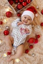 Cute smiling little girl in Santa Claus red hat is playing with wooden toy on a beige plaid with Christmas decorations. Royalty Free Stock Photo