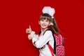 Cute smiling little girl in school uniform and white bows with backpack on red background. Back to school Royalty Free Stock Photo