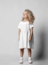 Cute smiling positive little blonde curly kid girl in white casual dress and sneakers is standing looking at upper corner