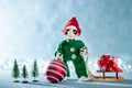 Cute Smiling Santas Helper Elf Holding Christmas Bauble and Pulling Christmas Gift on a Sledge. North Pole Christmas Scene.