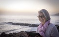Cute and smiling senior woman gray hair sitting near the beach surfing the net with tablet. Dusk light and horizon over water.