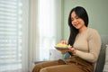 Cute smiling young Asian woman sitting on couch while eating a piece of cake. People, food and domestic life