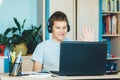 Cute smiling young boy in blue shirt sitting behind desk in his room next to laptop and study. Teenager in earphones waving hand