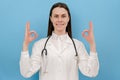 Cute smiling young female doctor in white coat with stethoscope showing ok sign and thumbs up, positive looking at camera, Royalty Free Stock Photo