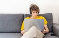 Cute smiling young teenager with laptop on the couch at home. Boy in white t shirt makes homework with a computer. Distance, Royalty Free Stock Photo