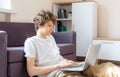 Cute smiling young teenager with laptop on the couch at home. Boy in white t shirt makes homework with a computer. Distance, Royalty Free Stock Photo