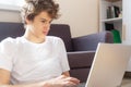 Cute smiling young teenager with laptop on the couch at home. Boy in white t shirt makes homework with a computer. Royalty Free Stock Photo