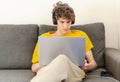 Cute smiling young teenager with laptop and headphones on the couch at home. Boy makes homework with a computer. Royalty Free Stock Photo