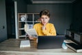 Cute student with a book in his hands sits at a table at home near a laptop and studies, looking at the camera with a surprised Royalty Free Stock Photo