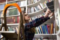 Pretty student stands on a stepladder in the library to find the right book. Woman in public library