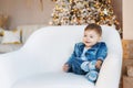 Cute stylish Caucasian baby boy in a blue denim suit and jacket poses sitting in a white chair. Christmas tree and gift Royalty Free Stock Photo