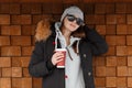 Cute stylish young hipster woman in a knitted hat in sunglasses in a black winter jacket with a fur hood in a stylish sweatshirt Royalty Free Stock Photo
