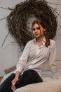 Cute stylish young woman in a fashionable white shirt in vintage black jeans enjoys a rest sitting on the bed in a cozy bedroom. Royalty Free Stock Photo