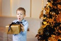 A cute surprised boy in a blue shirt near the Christmas tree with a gift in a gold package stretches forward. Selective focus Royalty Free Stock Photo