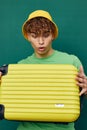 a cute, surprised man stands on a green background, dressed in a yellow panama hat, holding a bright travel suitcase in Royalty Free Stock Photo