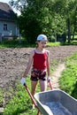 cute teenage girl in baseball cap and shorts pushing large garden wheelbarrow. Work on garden plot, in village on farm. Royalty Free Stock Photo