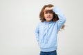 Cute teenage girl in a blue hoodie seeks out while holding a hand near her forehead on a light gray background