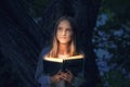 A cute teenage girl with long blonde hair is reading a magic book with light rays coming out from the inside, in a forest near a Royalty Free Stock Photo