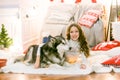 A cute teenage girl with long dark curly hair in a white pullover in a room with a Christmas decor with a large Malamute.