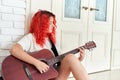 Cute teenage girl with red hair sits on the floor and plays a red guitar