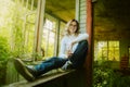 A cute teenager with blonde hair and jeans is sitting on the windowsill of a wooden abandoned house on a sunny summer day Royalty Free Stock Photo