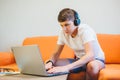 Cute teenager in white t shirt sitting on couch next to laptop and study. Serious boy in headphones makes homework,